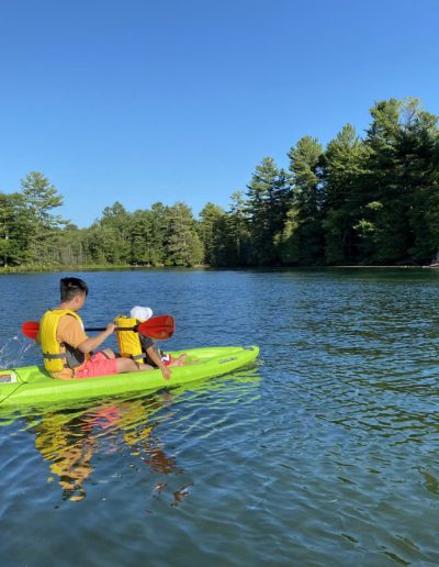 Two people in yellow life jackets paddling a green kayak on a lake surrounded by trees.