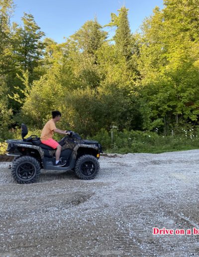Person in orange shirt and pink shorts riding an ATV on a gravel path near green trees.