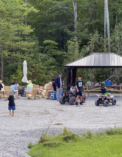 Outdoor gathering with people seated by a fire pit and children riding ATVs near a shelter.
