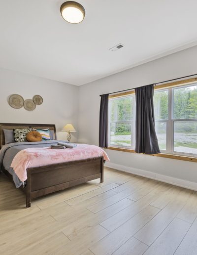 Spacious bedroom with wooden bed, nightstands, lamps, large window with dark curtains, and wood flooring.
