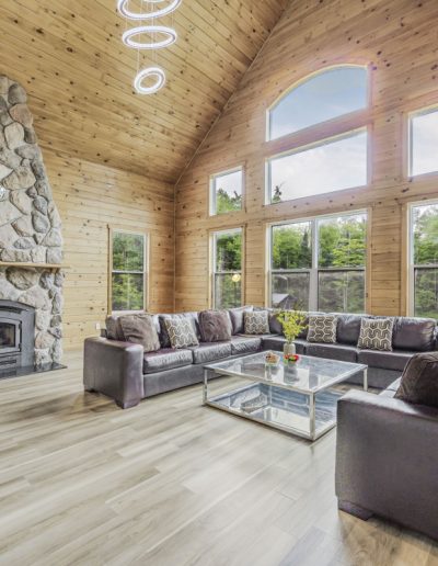 Living room with vaulted wood ceiling, stone fireplace, leather sectional sofa, and large windows.