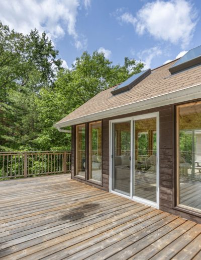 Wooden deck with railing adjacent to house with sliding glass doors and skylights, surrounded by trees.