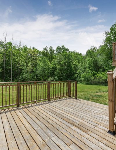 Wooden deck with railings and stairs overlooking grassy yard and dense trees under blue sky