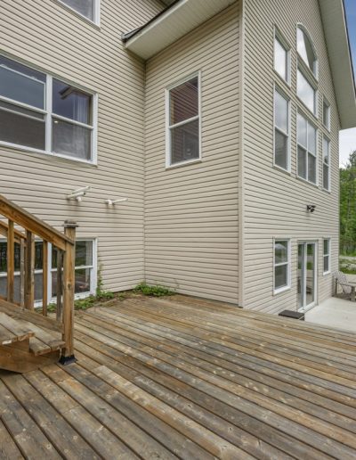 Wooden deck and stairs attached to beige house, concrete patio with chair, lawn, and trees.