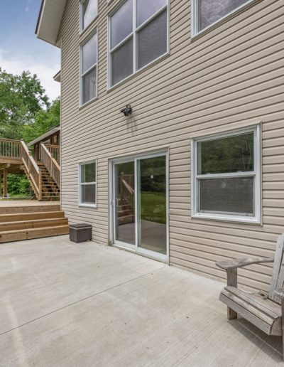 Beige house exterior with sliding glass door, concrete patio, wooden chair, stairs, and green trees.