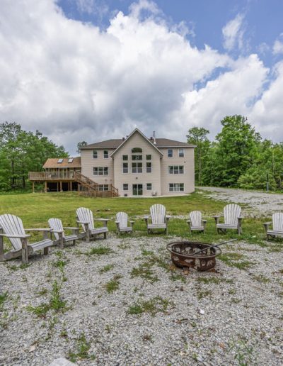 Two-story house with deck surrounded by trees, grassy area, and fire pit with chairs.