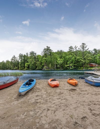 Kayaks, canoe, and paddle boat on sandy shore by lake with dock and forested background.