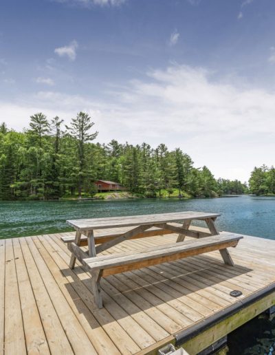 Wooden dock with picnic table on a lake surrounded by green trees and a cabin nearby.