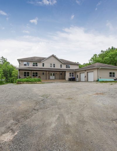 Two-story beige house with attached garage, covered porch, gravel driveway, and surrounding trees.