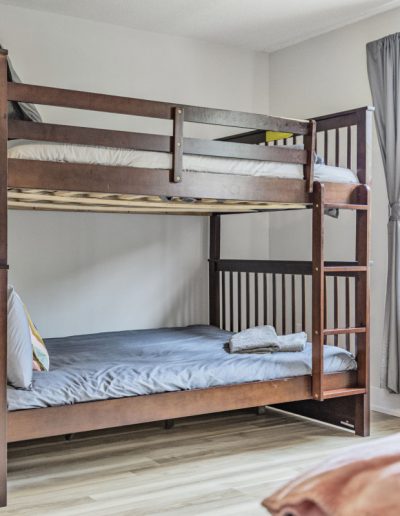 Wooden bunk bed with gray and white bedding, folded towels, beige chair, window with gray curtains.