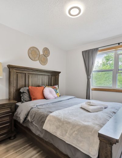 Bedroom with wooden bed and nightstand, gray bedding, colorful pillows, towels, window with gray curtains.