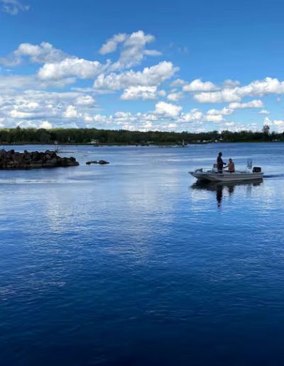 Small motorboat with two people on calm water near rocky shore and distant tree-covered land.