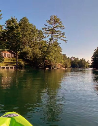View from green kayak on calm water toward tree-lined shore with wooden cabin and dock.