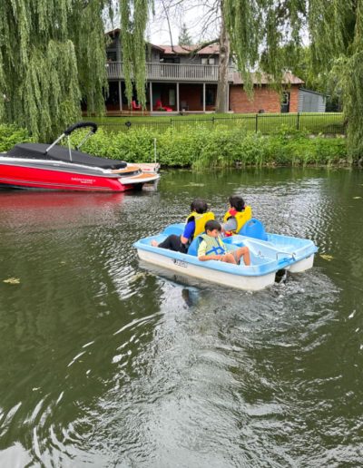 Three children wearing life jackets paddle a blue and white boat on a lake near a red motorboat and a house.
