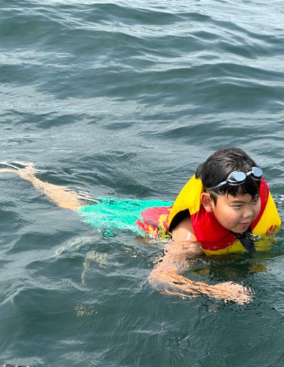 Child wearing a yellow and red life jacket and swim goggles swimming in open water.