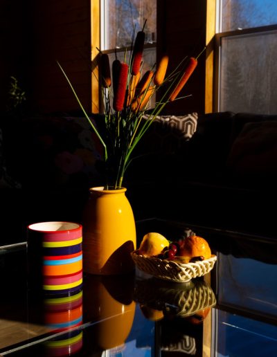 Glass-top coffee table with yellow vase, cattail plants, fruit basket, and striped mug by windows.
