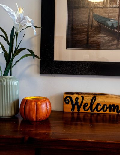 Wooden table with a green vase of white flowers, orange candle holder, welcome sign, and wall art.